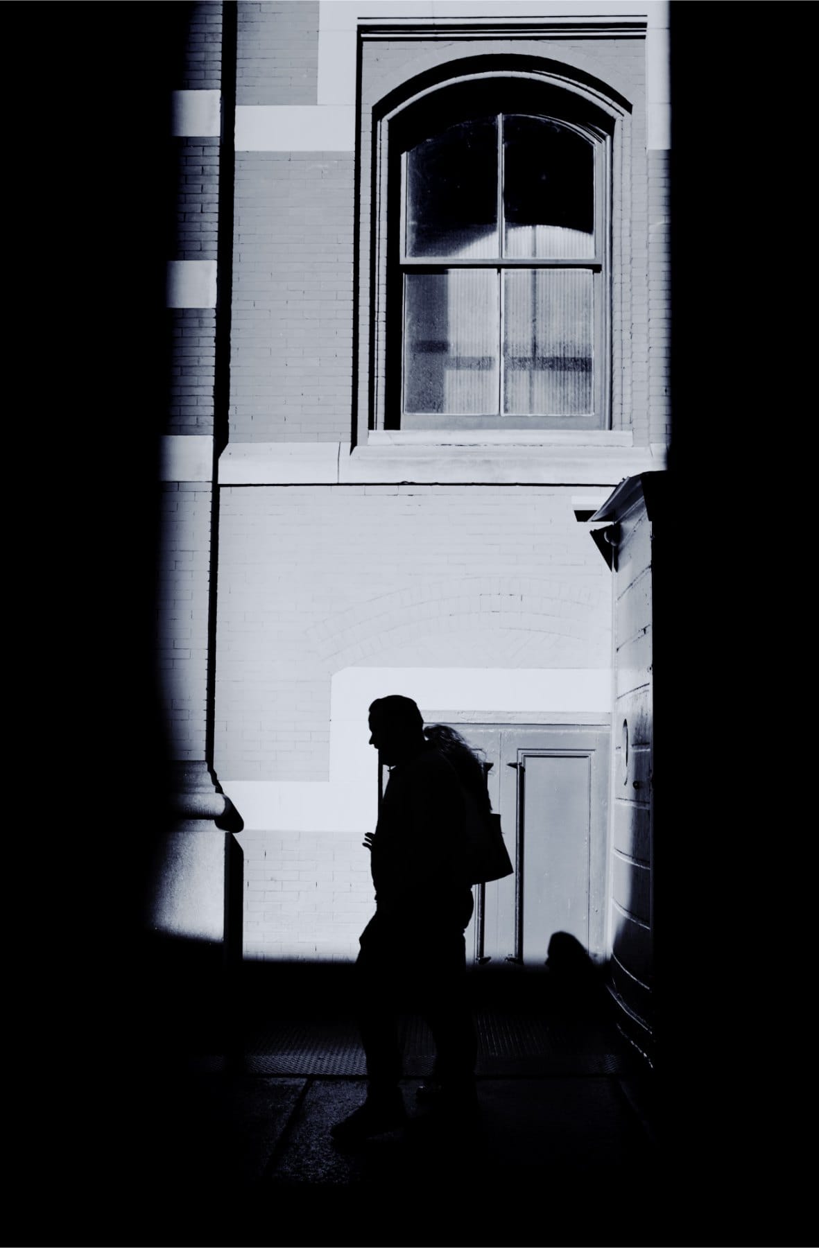 Black-and-white view through a dark doorway to a sunlit wall with an arched window and a silhouetted man with a backpack.