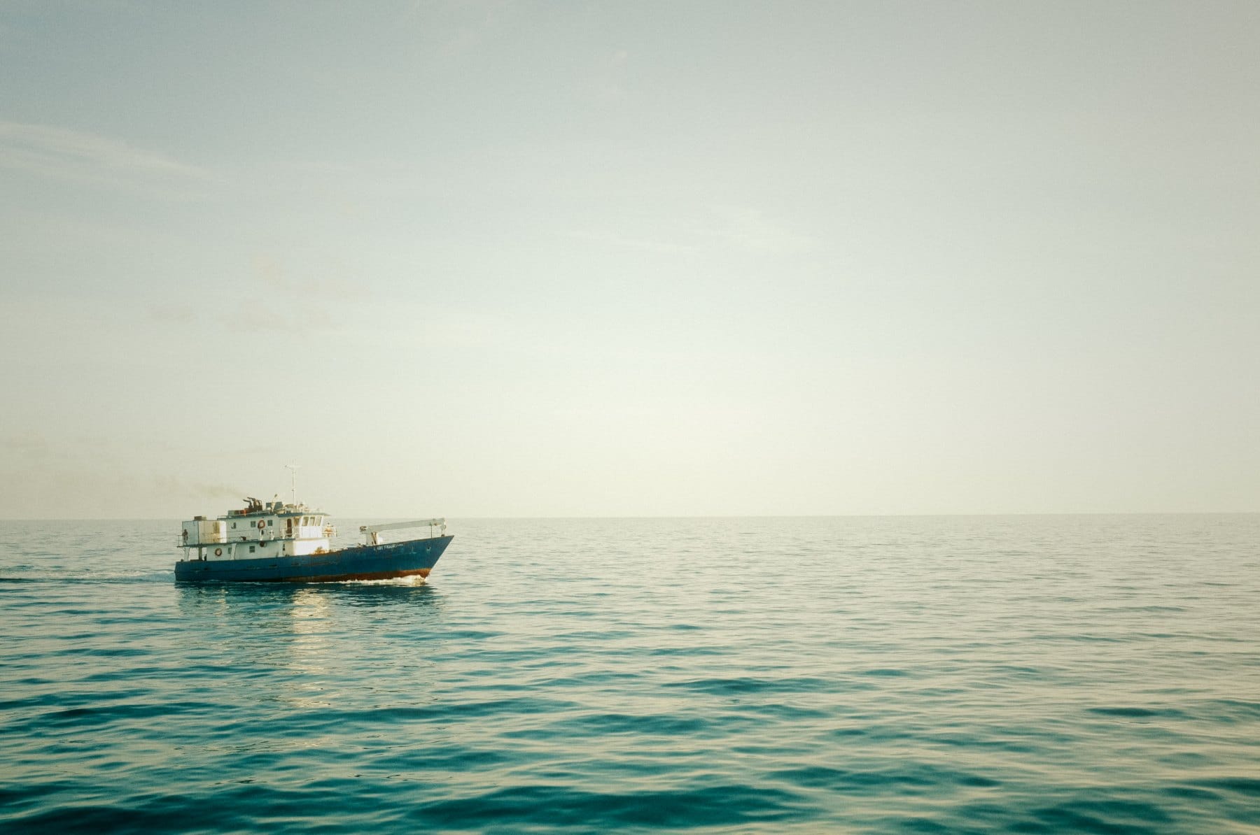 Blue-and-white work boat alone on calm teal water under a pale, hazy sky with a low horizon.