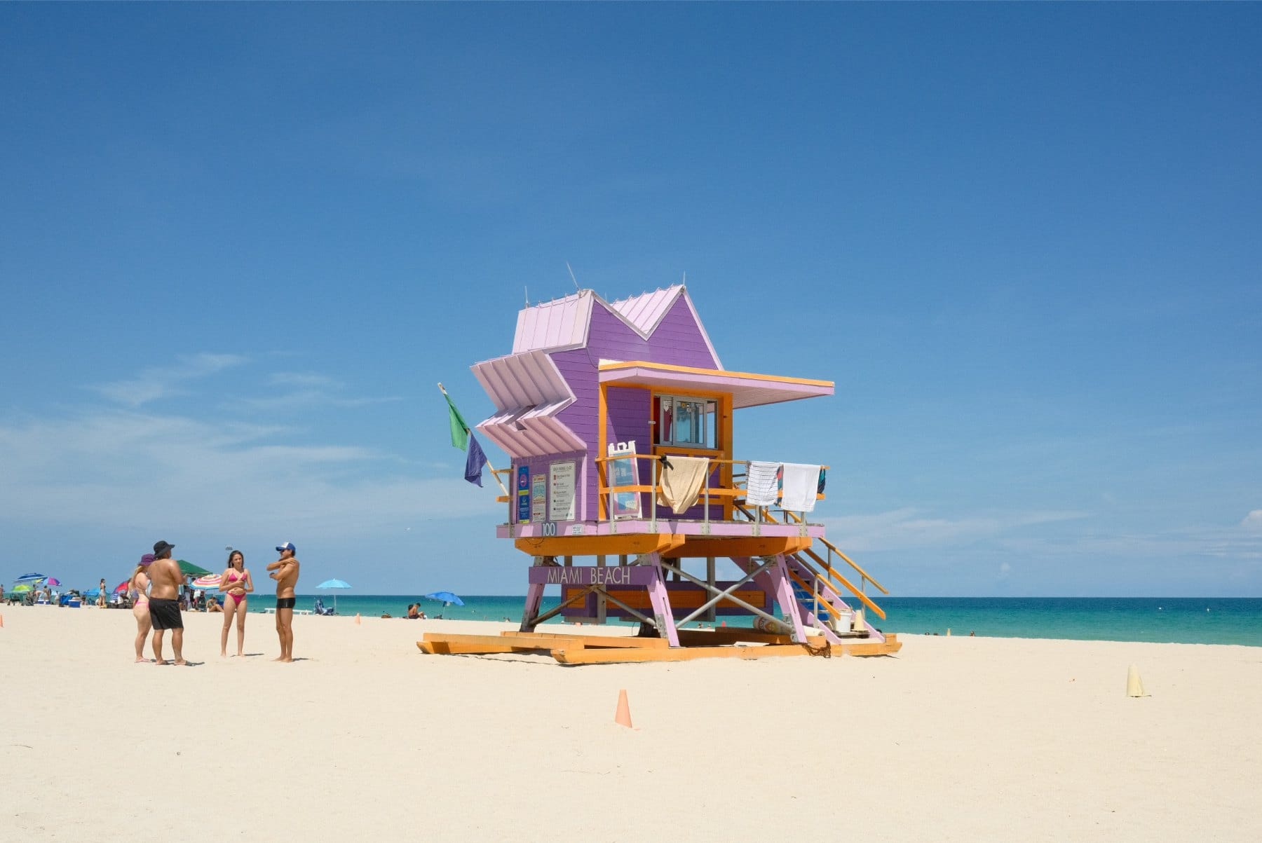 Purple and orange lifeguard tower on white sand under blue sky, with beachgoers and turquoise sea nearby.