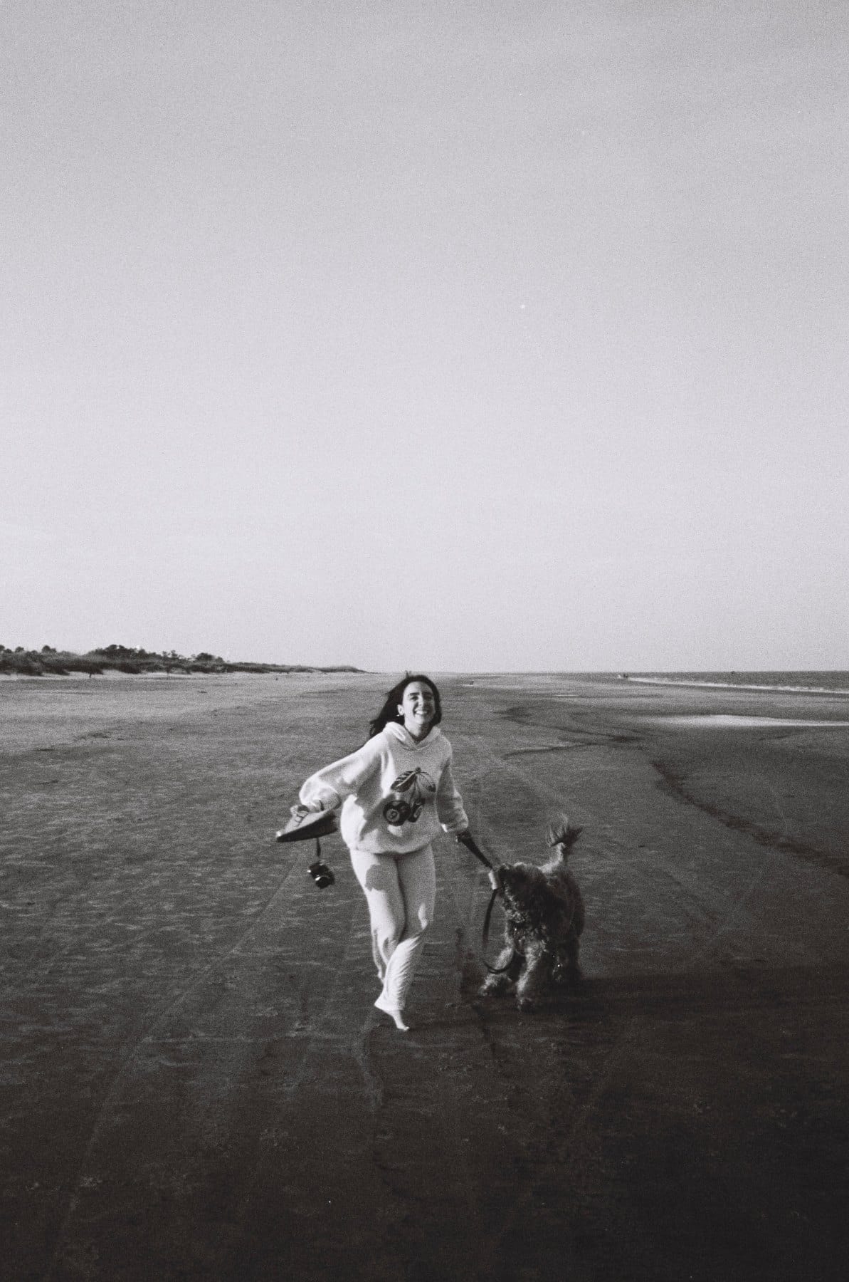 Black-and-white film photo of a woman laughing as she walks a curly-haired dog across wet sand on a wide beach.