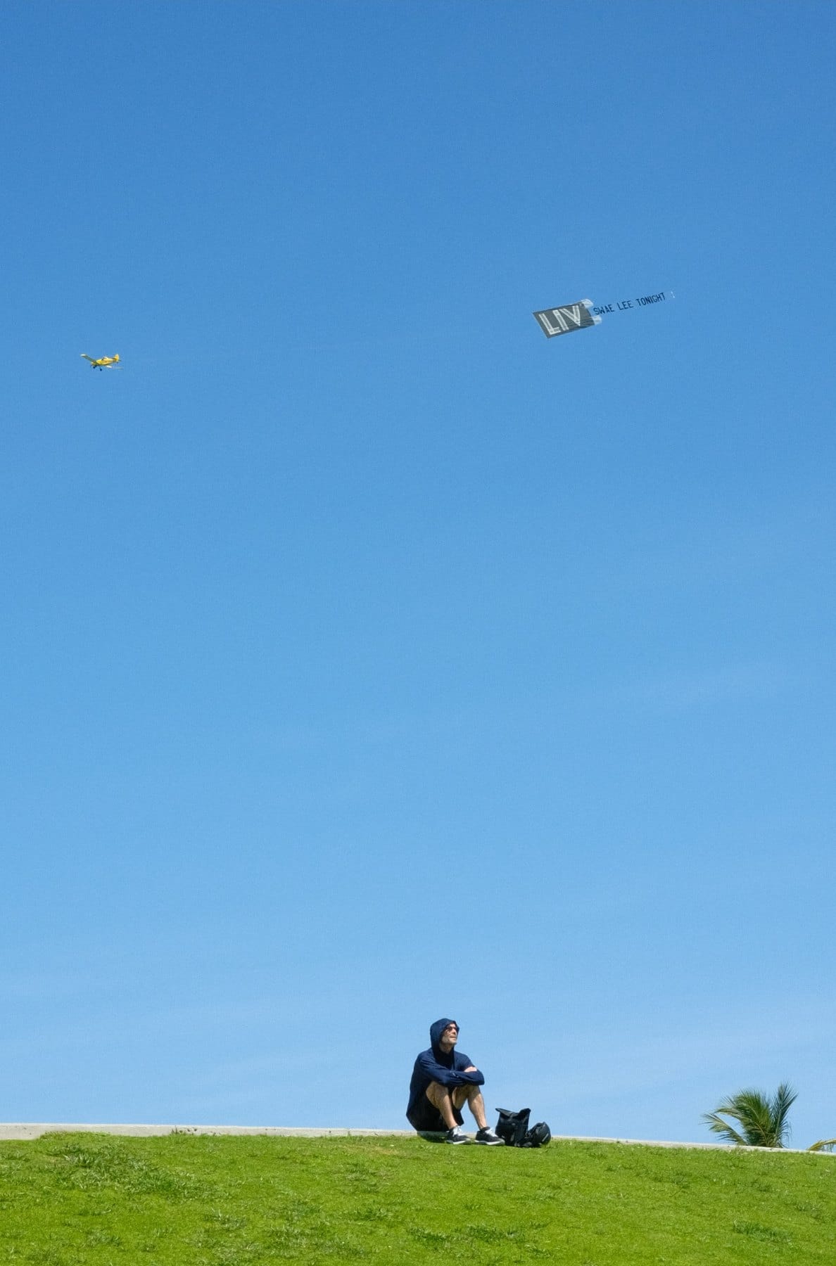Person sitting on a green hilltop beneath a vast clear blue sky, with two small kites flying high above.