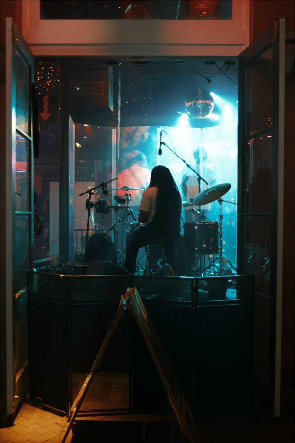Long-haired drummer behind a kit in a glass-walled stage booth lit in teal, seen through open doors.