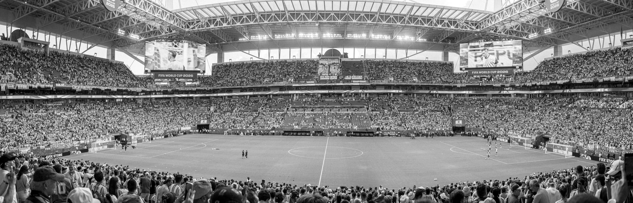 Wide panorama of a packed football stadium in black and white, with the green pitch and screens in color.
