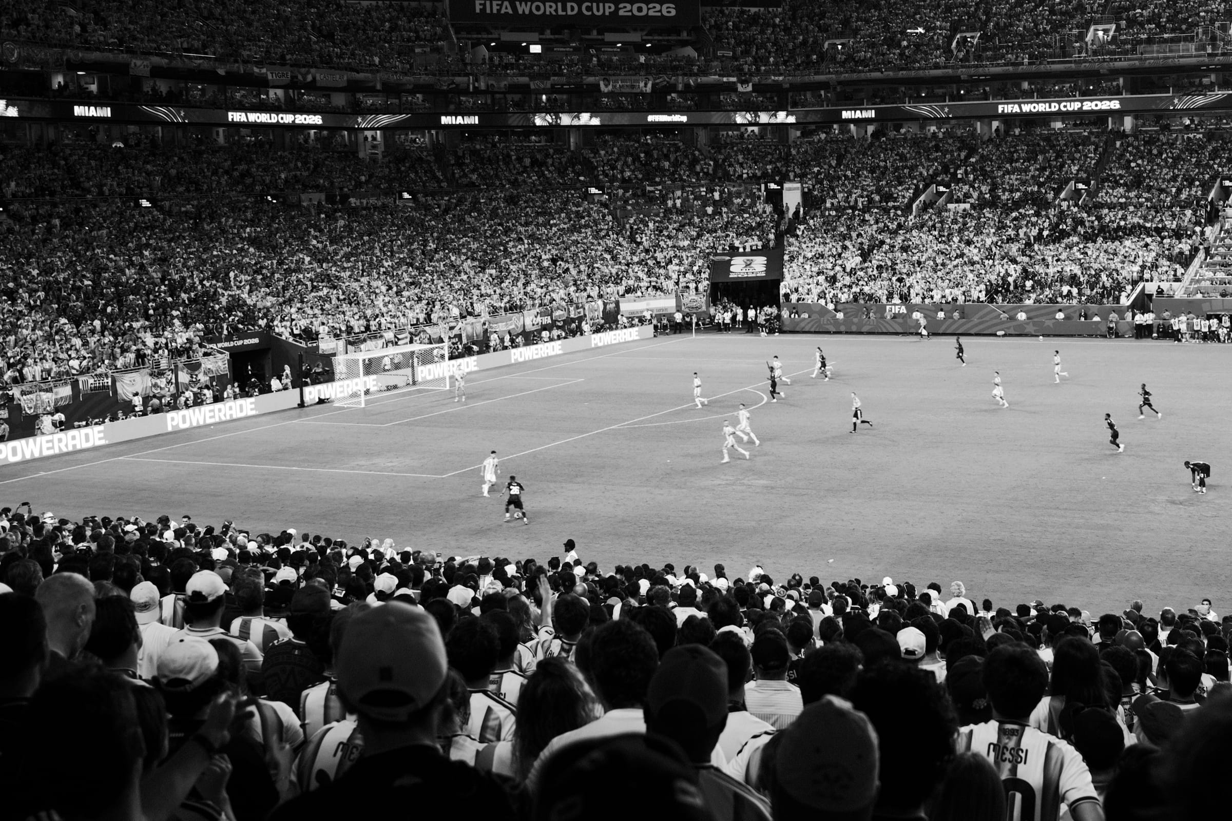 Black-and-white view over a crowd of fans toward players on the pitch at a FIFA World Cup 2026 match.