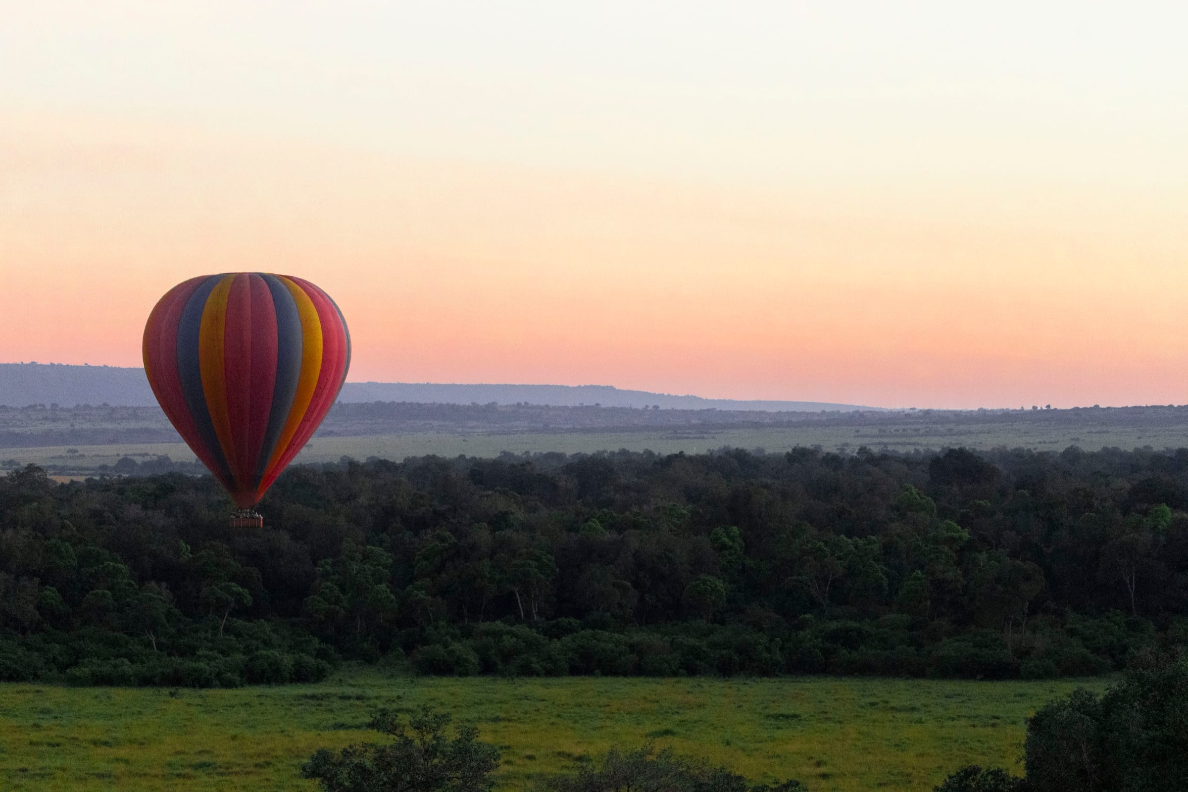 Striped hot-air balloon floating above a dark band of forest under a pink and peach dawn sky.