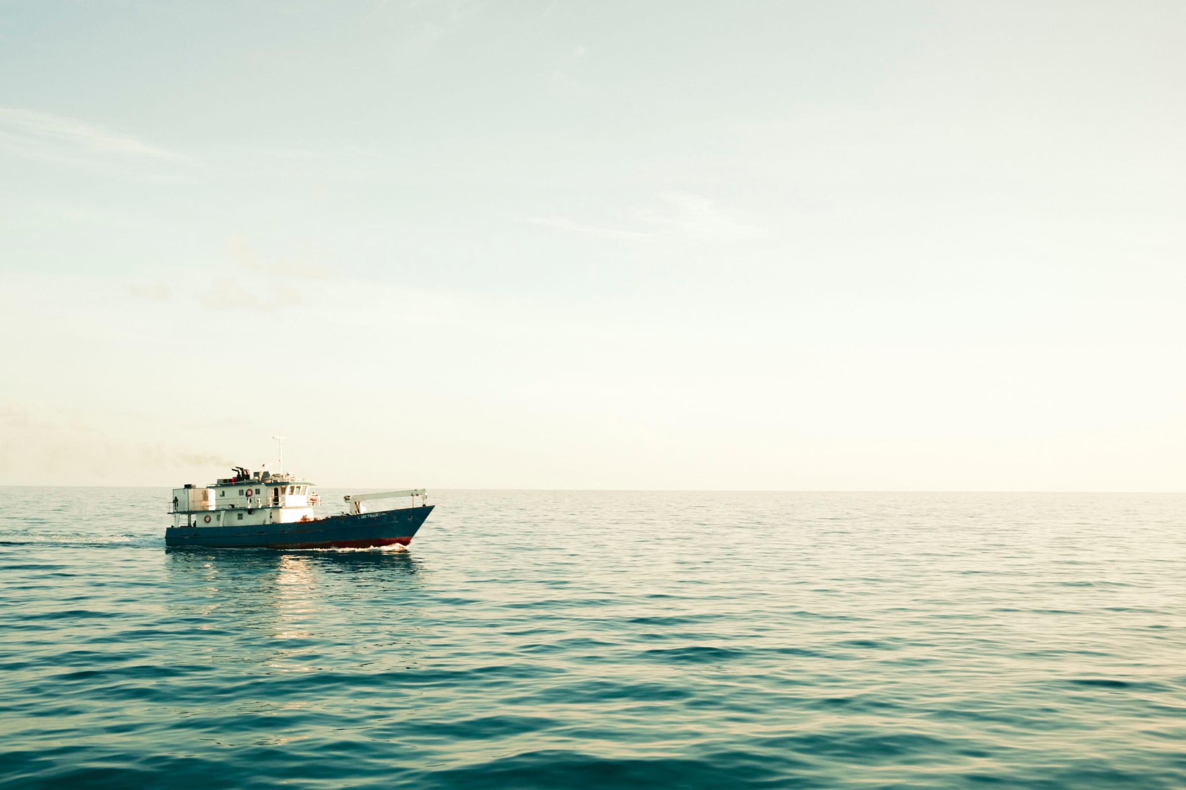 Blue-and-white work boat crossing calm teal sea under a soft, pale sky.