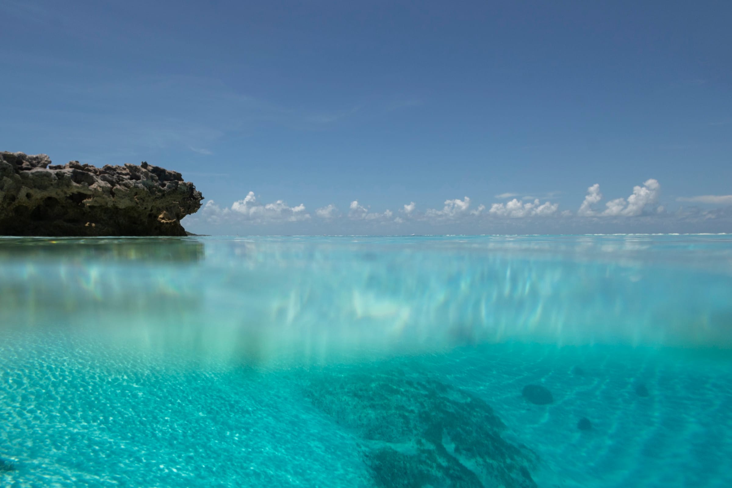 Split-level shot of a rocky headland and clouds above the waterline, with clear turquoise water and sand below.