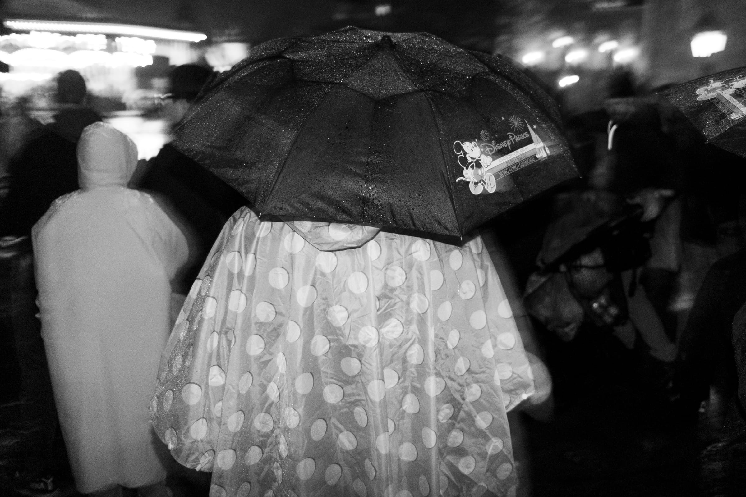 Black-and-white photo of crowds in ponchos with an umbrella in the rain, with lights blurred behind them.