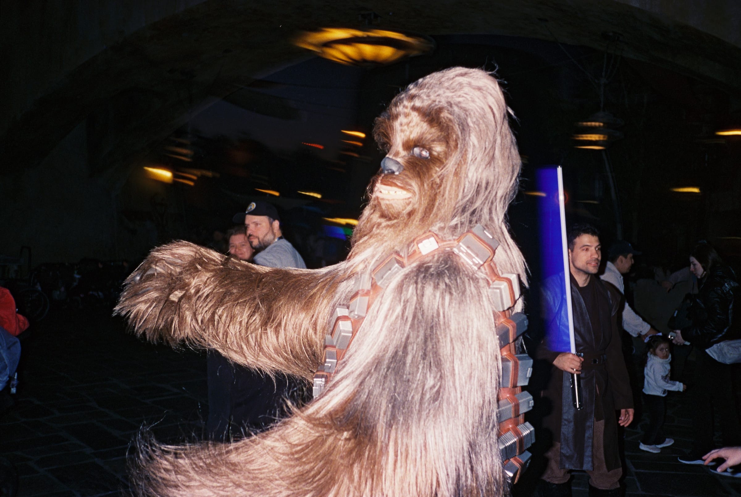 Hard-flash photo of a Chewbacca character reaching out in a dark hall, with a guest holding a blue lightsaber.