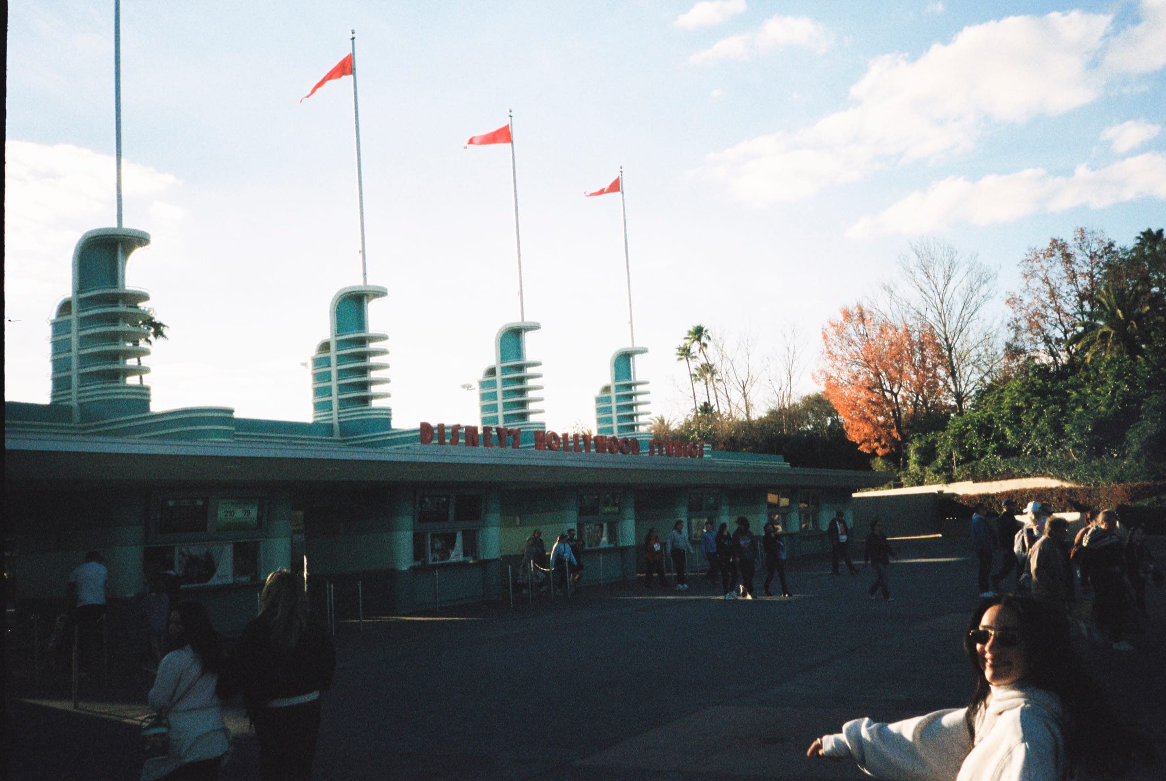 Film photo of an art-deco entrance with finned towers and red flags in low sun, a smiling friend in the corner.
