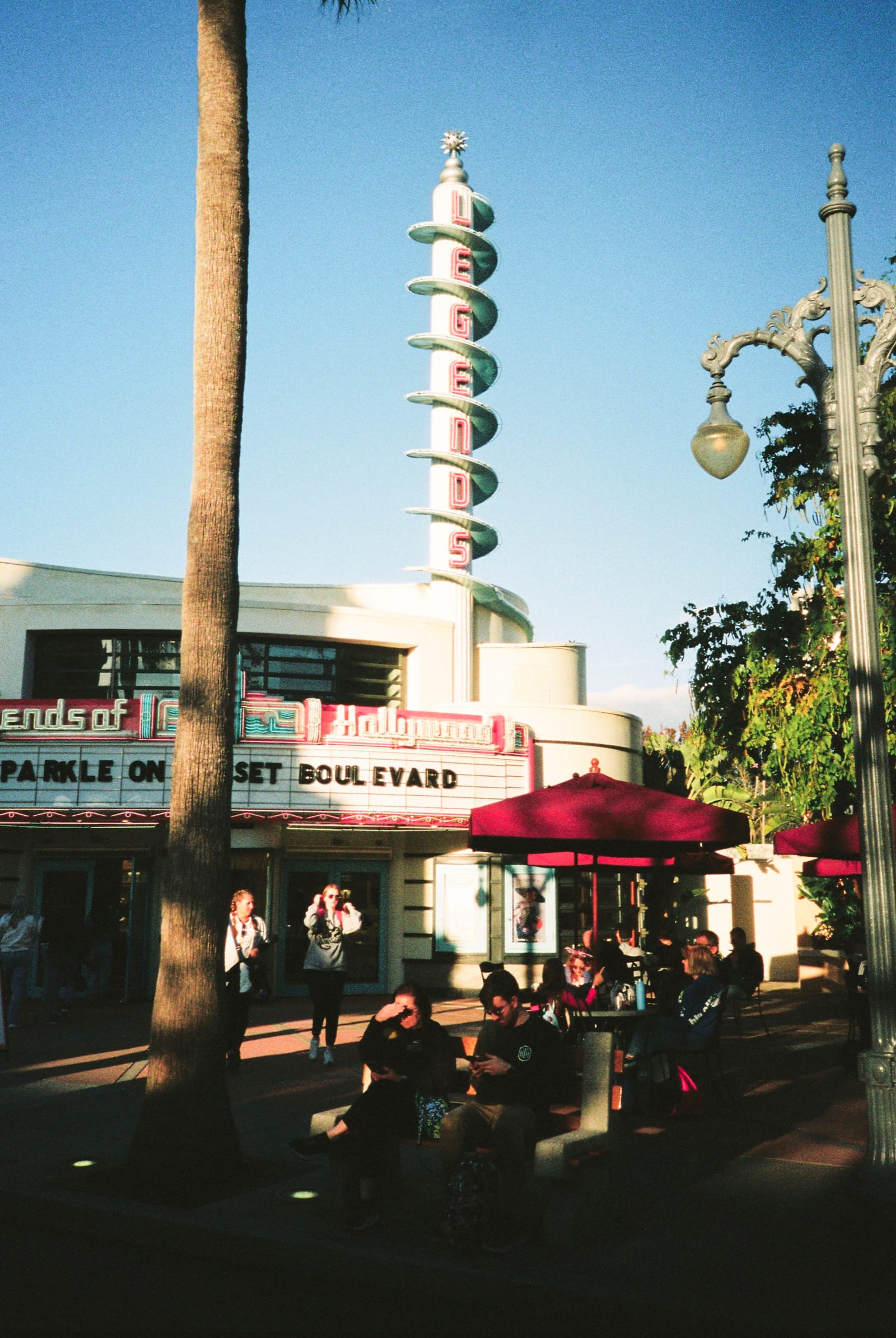 Film photo of an art-deco tower and marquee on a sunny street, with palms, red umbrellas and guests.