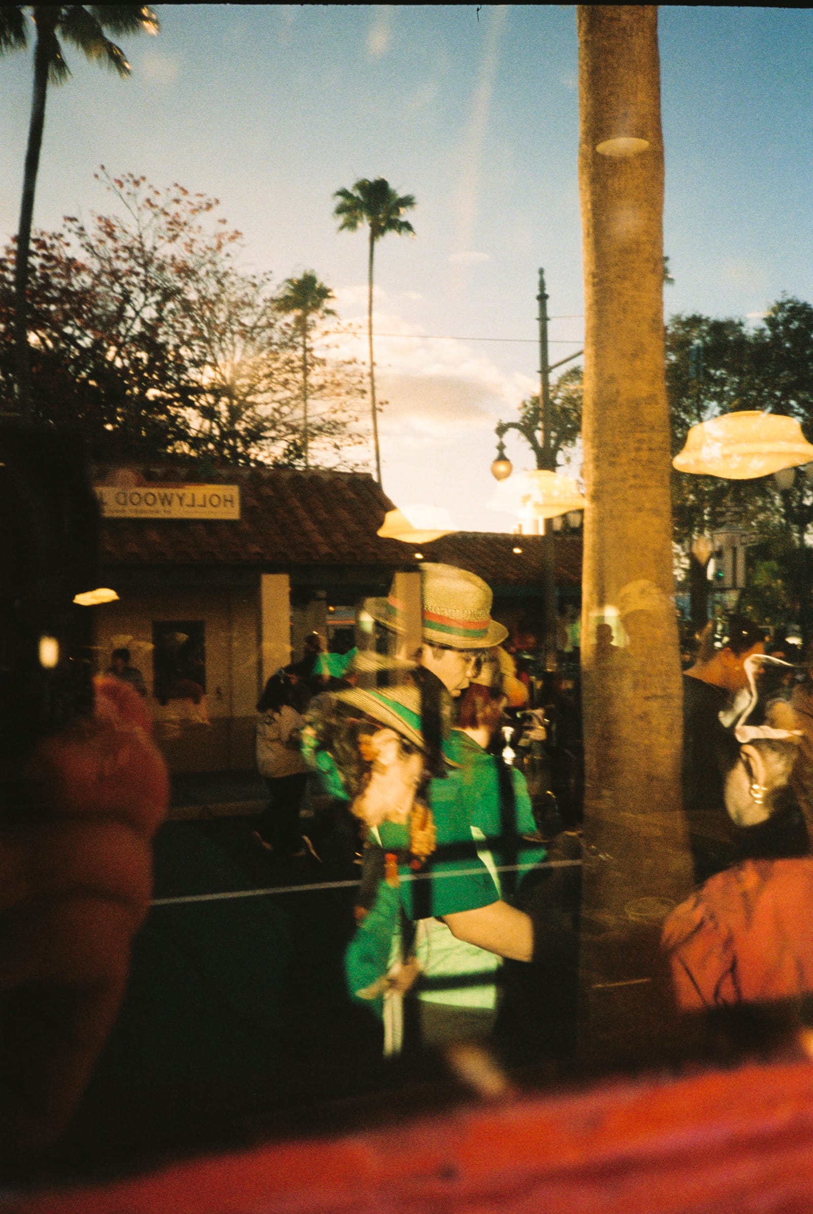 Film photo through glass blending reflected palms and buildings with a costumed figure in green inside.