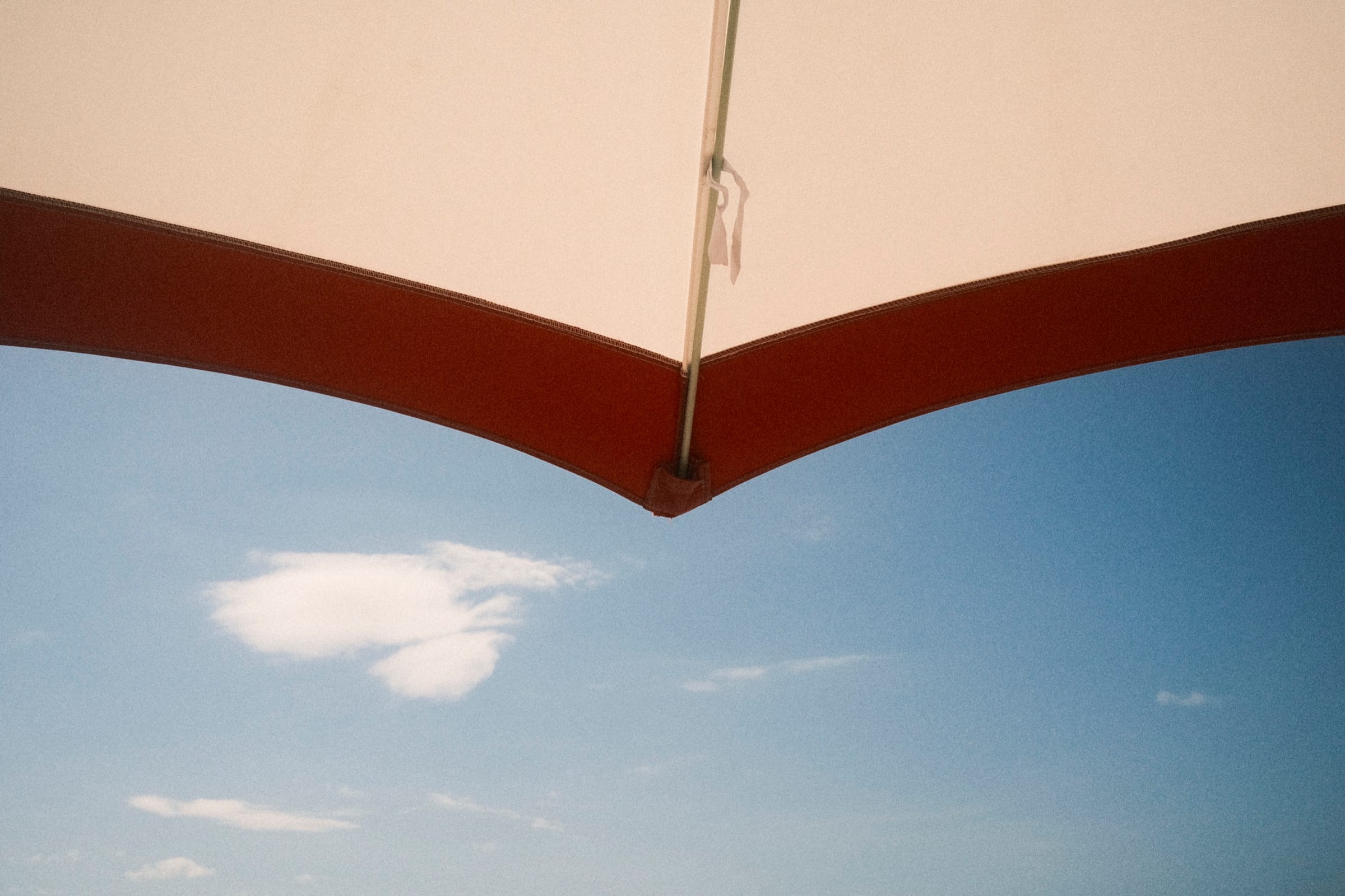 View from beneath the edge of a cream-and-red umbrella to blue sky and a single soft cloud.