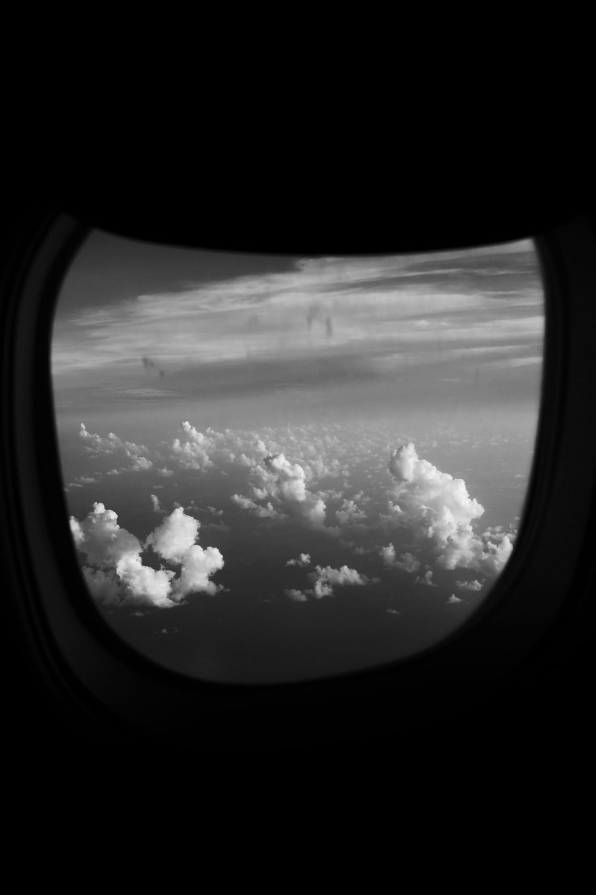 Black-and-white photo of puffy clouds seen through an airplane window, framed by the dark cabin wall.